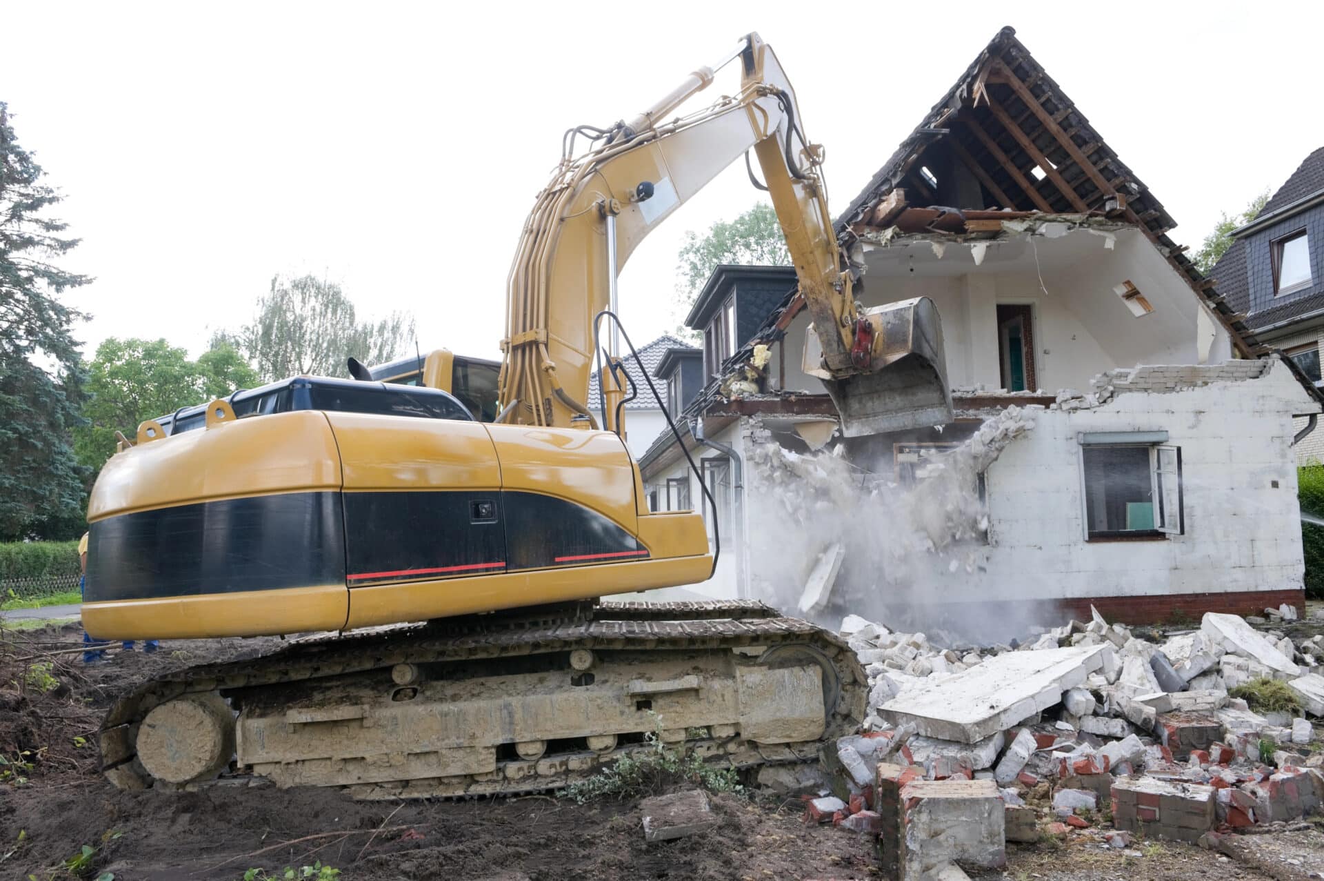 Excavator demolishing a damaged house during water damage recovery, with debris scattered around the site.