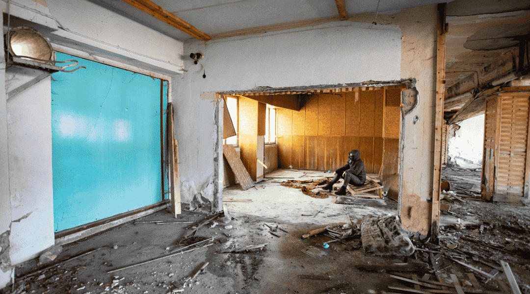 Interior of a damaged room with debris, showing a person seated on wooden planks, emphasizing the impact of water damage and the need for restoration services.